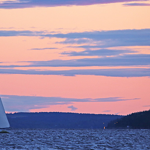 A sailboat cuts across Kennebecasis Bay with the Kingston Peninsula and Westfield ferry docking lights visible in the…