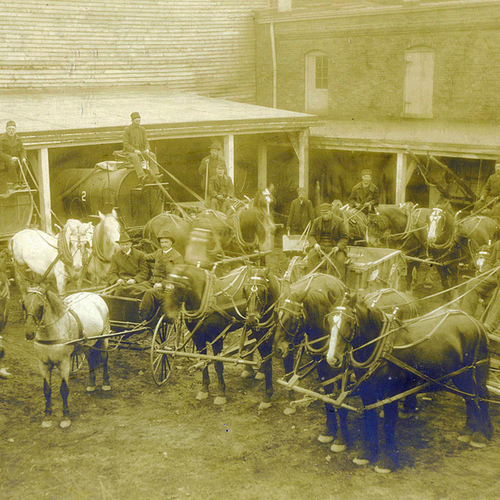 A large group courtyard photo from the last quarter of the 19th century showing horse drawn water tankers, wheel barr…