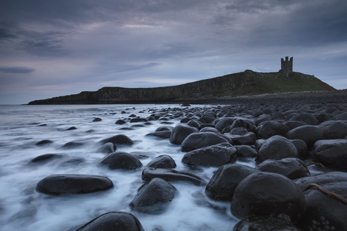 Dunstanburgh Castle at dawn as the cold North Sea moves in around the boulders.