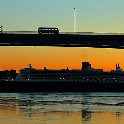 The Queen Mary 2 can be seen docked within Saint John Harbor as a double decker tour bus passes over the harbor bridg…