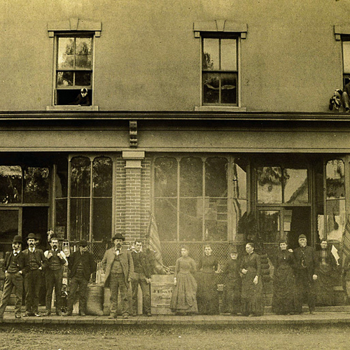 Employees in period dress stand outside a dry-goods store with others in the window above. A sign on a crate along th…