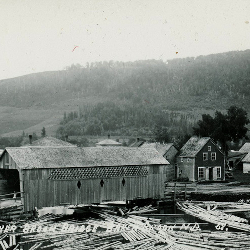 Logs float along and under Baker Brook Covered Bridge with vintage homes in the backdrop, in Madawaska County, New Br…