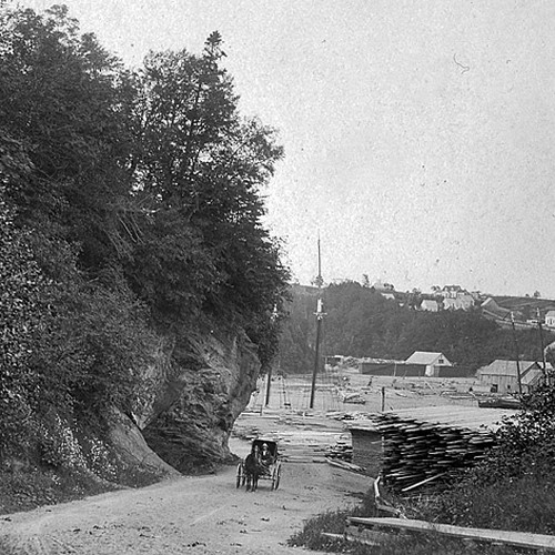 A horse & carriage pass a lumber stock and approach the camera's pov in an early 1900s view of St. Martins, NB, Canada